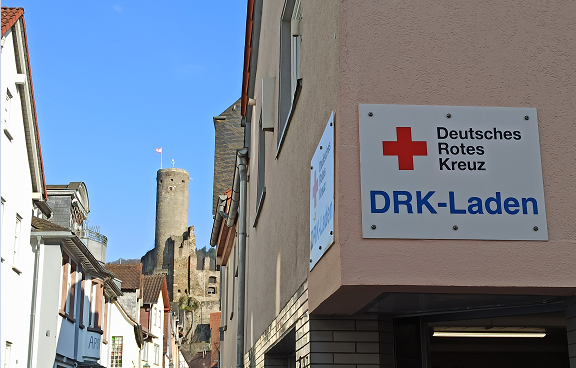 Das DRK-Laden-Schild ist im Vordergrund am Gebäude zu sehen. Im Hintergrund sind viele Gebäude, ein Turm und der blaue Himmel