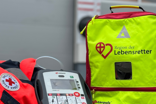 EH_Lebensretter_ReferenzBild Eine neongelbe Tasche des Projekts "Region der Lebensretter" steht neben einem Defibrillator.