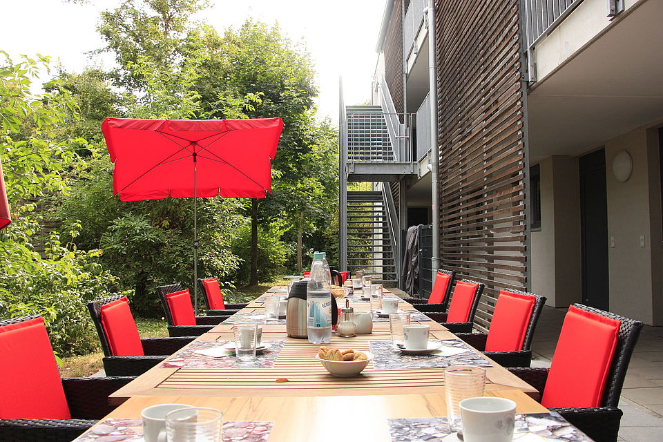 BW Terrasse Langer Esstisch auf einer Terrasse mit gedecktem Geschirr, roten Sitzkissen und großem Sonnenschirm, umgeben von Grün.