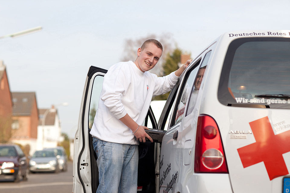 DRK - Menüservice Ein Mann im DRK-Pullover steht in der Tür eines weißen Fahrzeugs und lächelt in die Kamera.