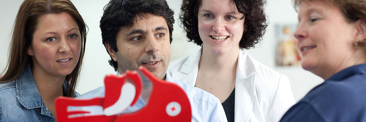 Kurse Erste Hilfe Erste Hilfe Kurs: Eine Mitarbeiterin des DRK steht seitlich zur Kamera und erklärt drei Personen etwas, die gespannt zuhören.