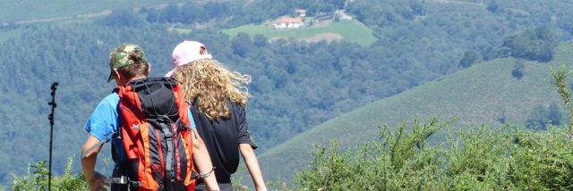 EH_Outdoor Ein Mann und eine Frau sind wandern und wurden von hinten fotografiert. Im Hintergrund sind Hügel, Bäume und ein Haus zu sehen.