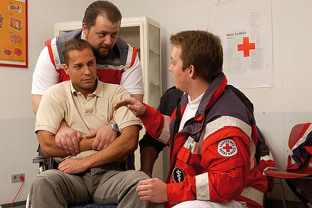 Rotkreuzkurs EH Fortbildung (BG) Ein Mann in DRK Uniform zeigt eine Hebetechnik anhand eines Mannes der sitzt. Ein anderer Mann in Uniform erklärt, wie es funktioniert.