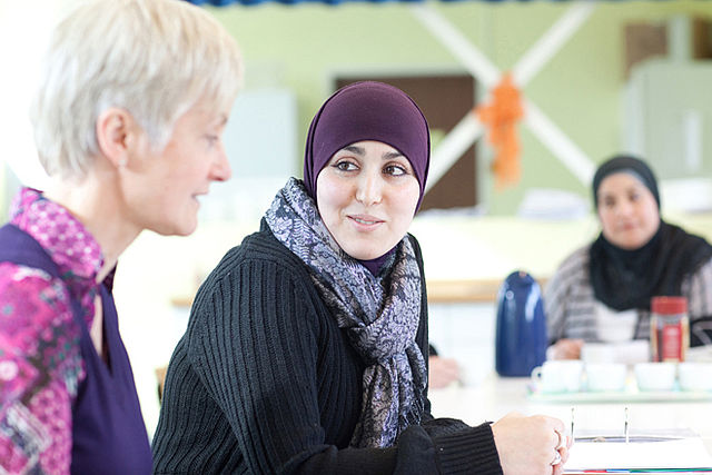 DRK - Beratung Migration, Mettmann Eine Frau mit lilanem Kopftuch unterhält sich mit einer älteren Dame. Im Hintergrund ist eine weitere Frau zu sehen.
