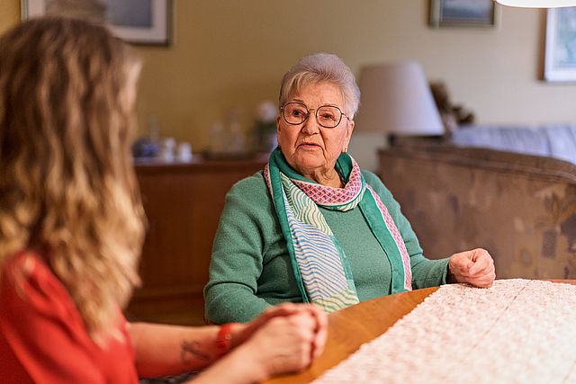 Seniorenberatung_resource Eine ältere Dame unterhält sich mit einer Frau, welche nur von der Seite verschwommen zu sehen ist. Sie sitzen in einem Wohnzimmer.