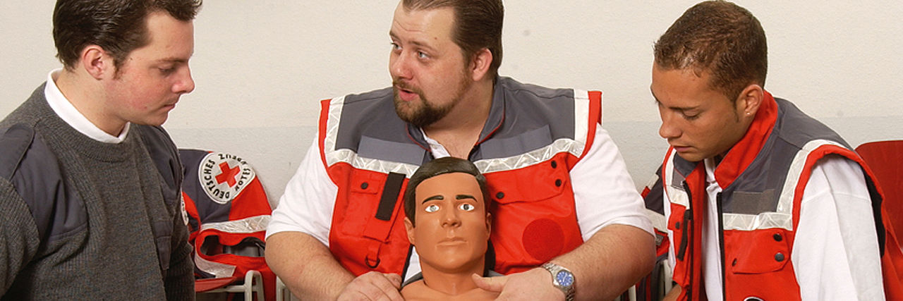 Rotkreuzkurs EH Fortbildung (BG) Erste Hilfe Kurs: Zwei Mitarbeiter des DRK knien vor einem Dummy und demonstrieren dem Zuschauenden die richtige Technik.