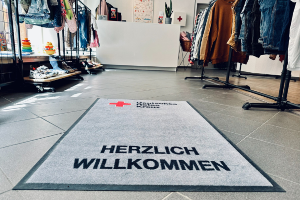 Kleiderladen Hochheim Innen Ein Teppich vom DRK liegt im Fokus, auf welchem "Herzlich Willkommen" steht. Am Rand sind die Füße der Kleiderstangen zu sehen.