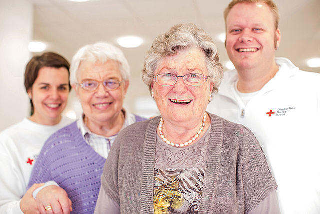DRK-Tages- und Begegnungsstaetten Zwei ältere Damen stehen lachend im Vordergrund. Hinter ihnen stehen eine Mitarbeiterin und ein Mitarbeiter mit Kleidung des DRK.