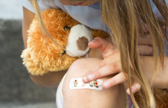Plasterkurs Content img Kind mit Pflaster am Knie und einem Teddy im Arm