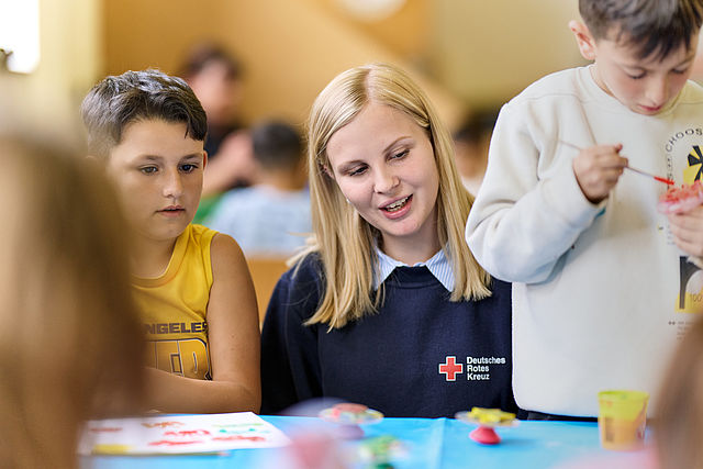FSJ_Content_Image Eine junge Frau in DRK Oberteil sitzt zwischen zwei Kindern und schaut diesen beim Basteln zu.