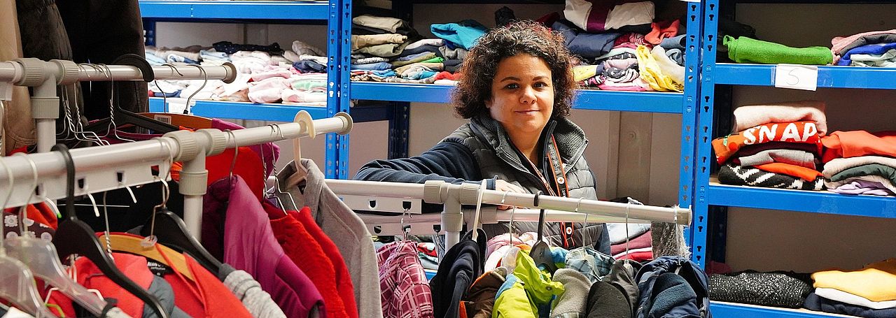 Kleiderkammer Hofheim Stage Image Eine Mitarbeiterin der Kleiderkammer steht inmitten von Kleiderstangen und Regalen mit Kleidung und lächelt in die Kamera.