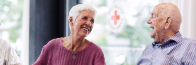 EH Fit für Senioren Ein älterer Herr und eine ältere Dame sind einander zugewandt und sprechen lächelnd miteinander. Im Hintergrund ist verschwommen eine Glasscheibe mit einem runden DRK-Aufkleber.