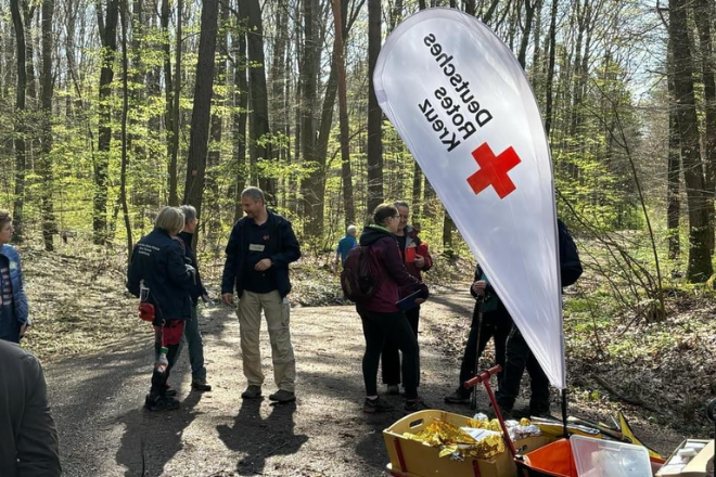 Die Teilnehmer stehen im Wald und unterhalten sich. Eine weiße Beachflag mit dem DRK Kreuz und der Aufschrift "Deutsches Rotes Kreuz" steht im Vordergrund.